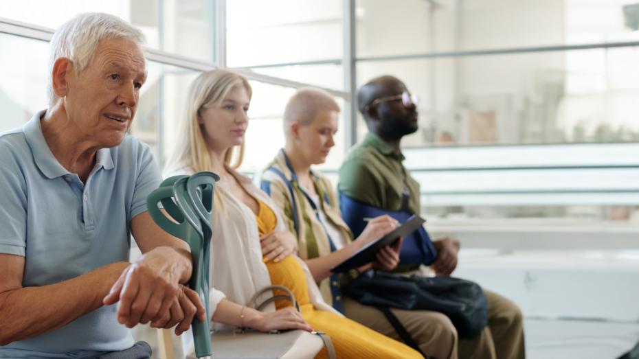 Vier Personen unterschiedlichen Alters und Aussehens sitzen nebeneinander in einem modernen Wartebereich. Ganz links sitzt ein älterer Mann mit grauen Haaren, der sich auf eine Gehhilfe stützt. Neben ihm sitzt eine schwangere Frau mit langen blonden Haaren. Rechts von ihr sitzt eine Frau mit sehr kurzen, möglicherweise durch eine Krankheit verlorenen Haaren, die ein Notizbuch oder Tablet hält. Ganz rechts sitzt ein Mann mit dunkler Hautfarbe, Bart und Brille, dessen rechter Arm im Gips ist. Im Hintergrund s